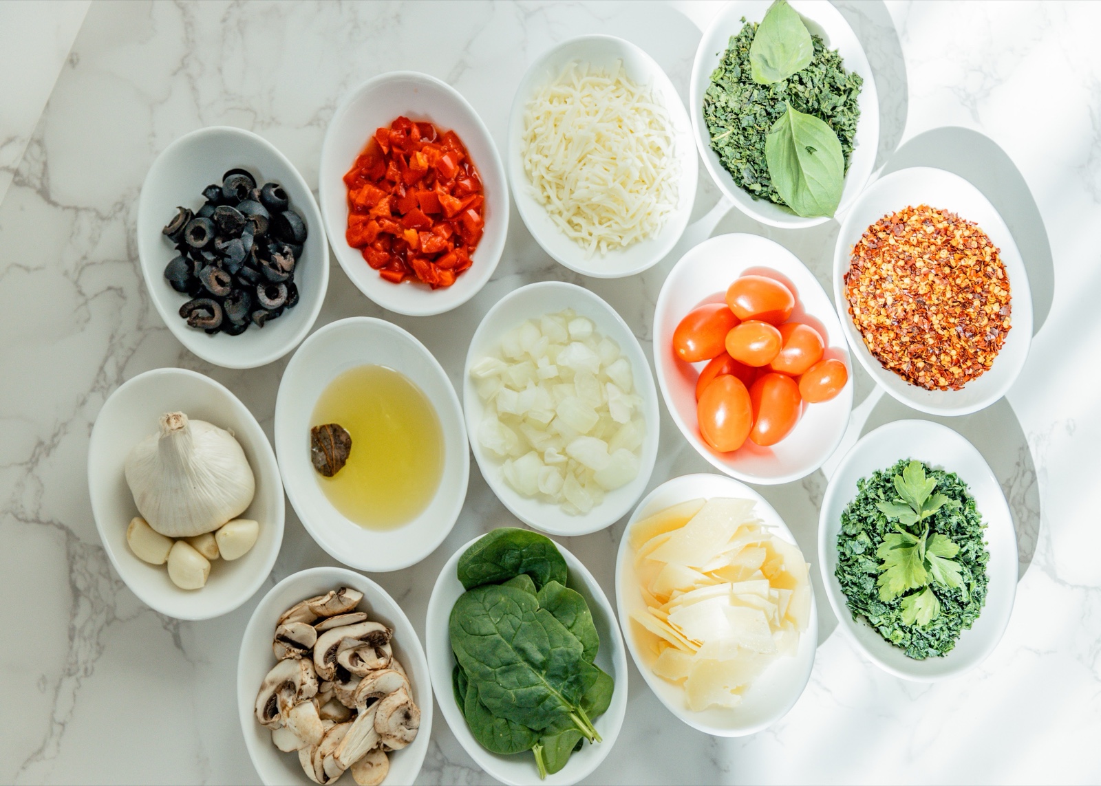 An overhead view of fresh Pastaphony ingredients on marble — olives, roasted peppers, parmesan, basil, garlic, cherry tomatoes, spinach, mushrooms